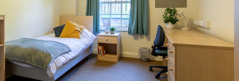 A student room in Churchill Hall, with a single bed in one corner and a desk opposite. A green space outside is visible through the window.